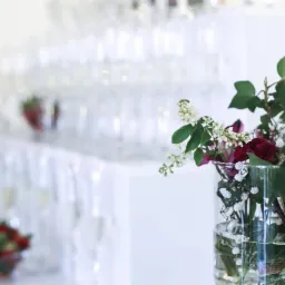 Dekoracja stołu z kwiatami w szklanym wazonie, w tle piramida z kieliszk&oacute;w do szampana i miseczki z owocami na białym obrusie, możliwe na imprezie integracyjnej.