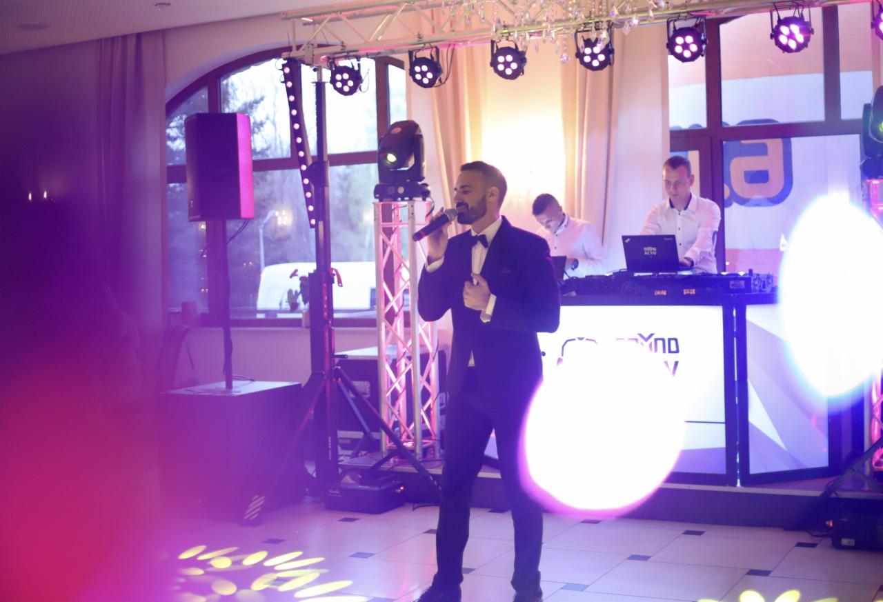 Wokalista w ciemnym garniturze i muszce śpiewa do mikrofonu na scenie podczas wydarzenia, za nim DJ przy konsolecie i oświetlenie sceniczne