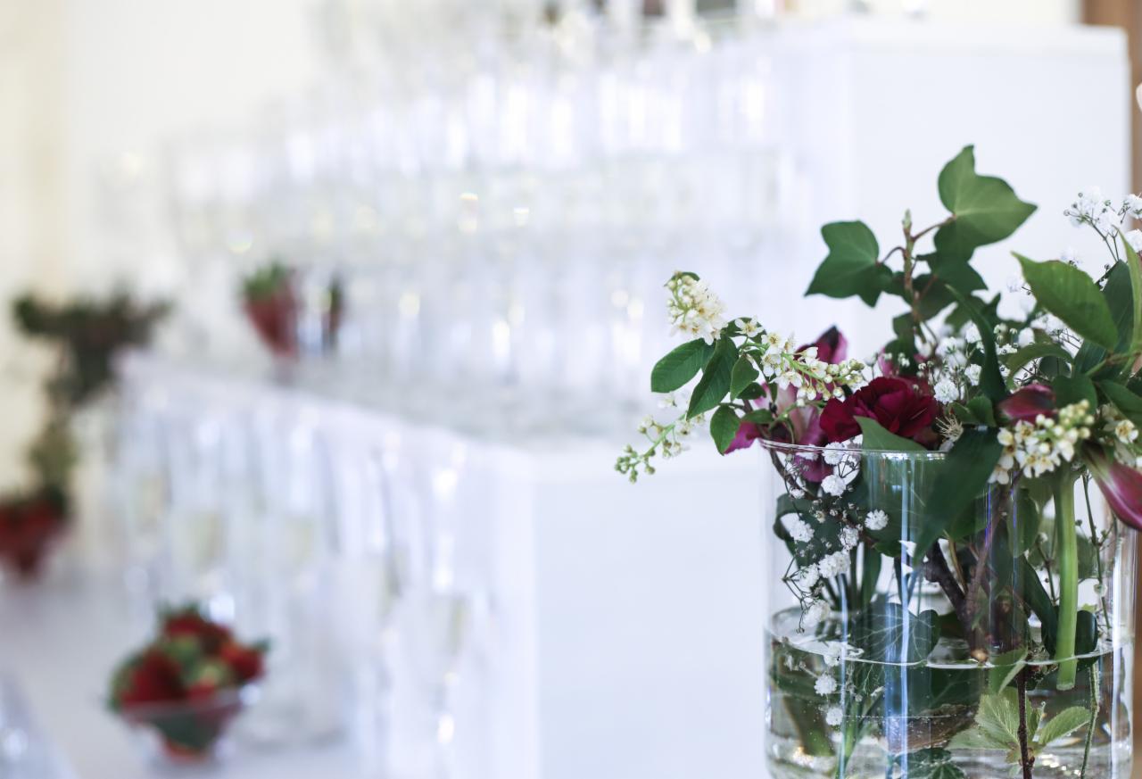 Dekoracja stołu z kwiatami w szklanym wazonie, w tle piramida z kieliszków do szampana i miseczki z owocami na białym obrusie, możliwe na imprezie integracyjnej.