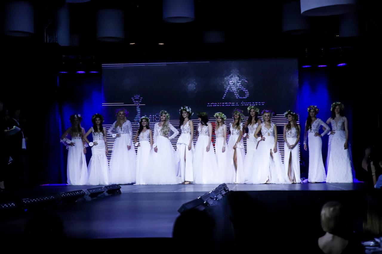 Modelki w białych sukniach z wiankami na głowach prezentują się na wybiegu podczas pokazu mody, oświetlonym niebieskim światłem, z dużym ekranem w tle.