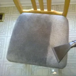 Fragment krzesła z szarą tapicerką w trakcie czyszczenia parowego, widoczna różnica w kolorze pomiędzy wyczyszczoną a nie wyczyszczoną częścią siedziska, na podłodze widoczne ślady wody.