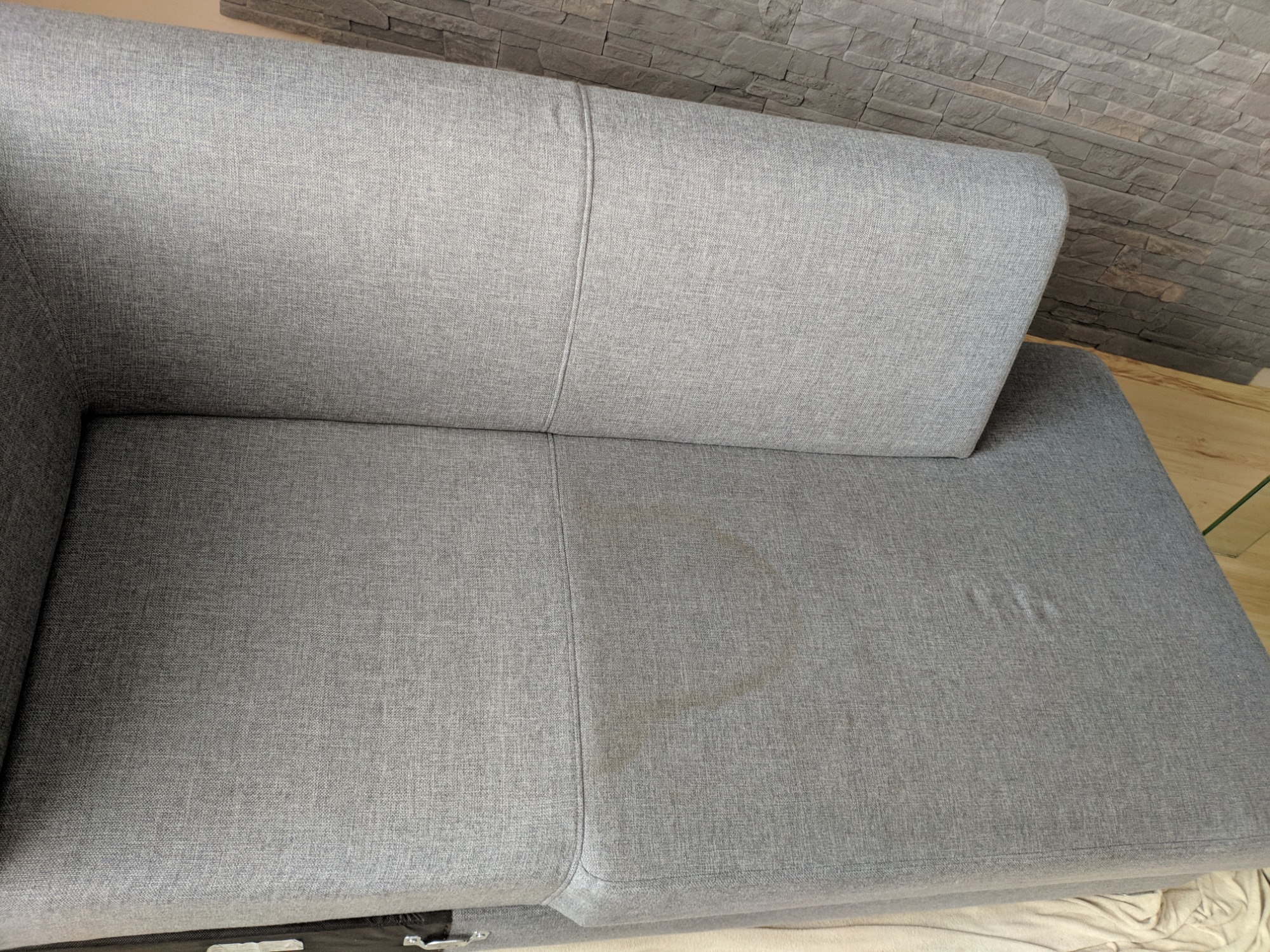 Szara kanapa z wyraźną plamą na siedzisku, widoczna faktura materiału i fragment ściany z dekoracyjnej cegły w tle.