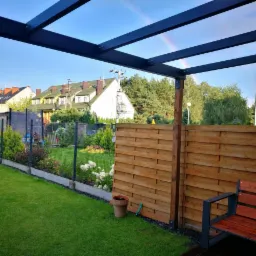 Nowoczesny ogród z drewnianym ażurowym ogrodzeniem, siatką ogrodzeniową, pergolą i tęczą w tle.