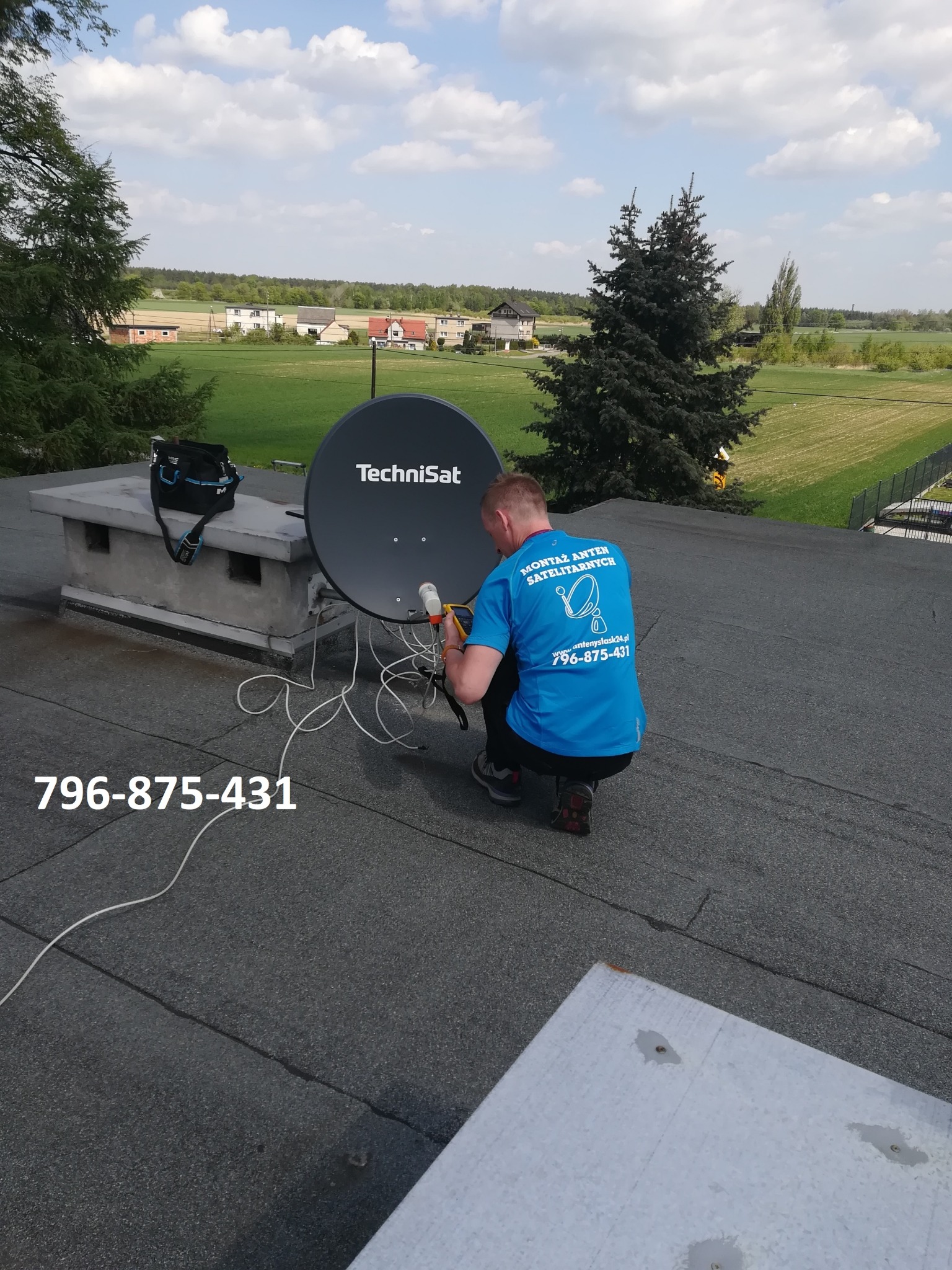 Instalator w niebieskiej koszulce z logo firmy, klęczy na dachu z papy i podłącza przewody do anteny satelitarnej TechniSat. W tle widoczny krajobraz z polami i drzewami. Numer telefonu firmy...