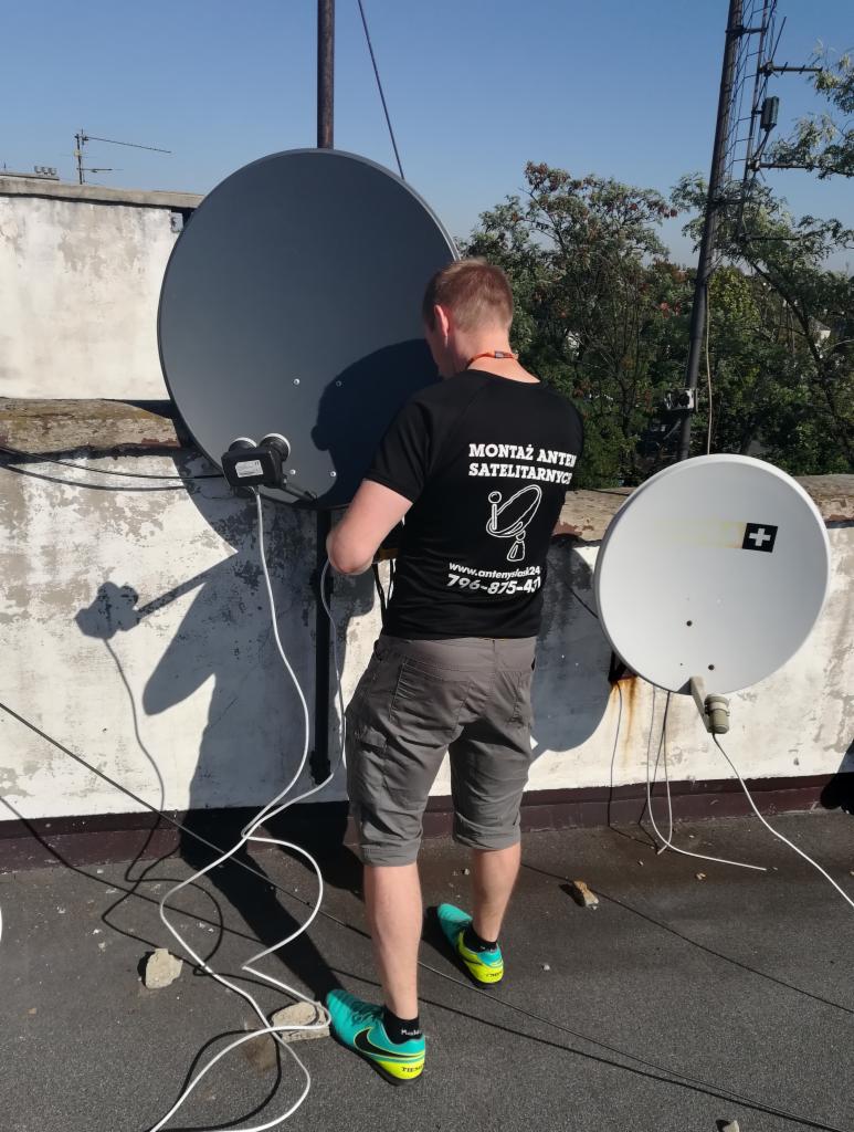 Instalator w czarnej koszulce z logo firmy, podłącza kable do szarej anteny satelitarnej na dachu budynku, obok widoczna biała antena.