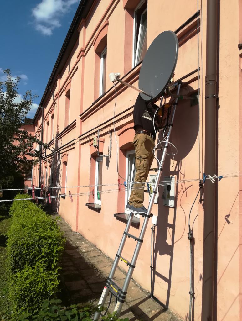 Pracownik na drabinie montuje talerz antenowy na elewacji budynku, widoczne kable i osprzęt, z boku krzewy, w tle okna.