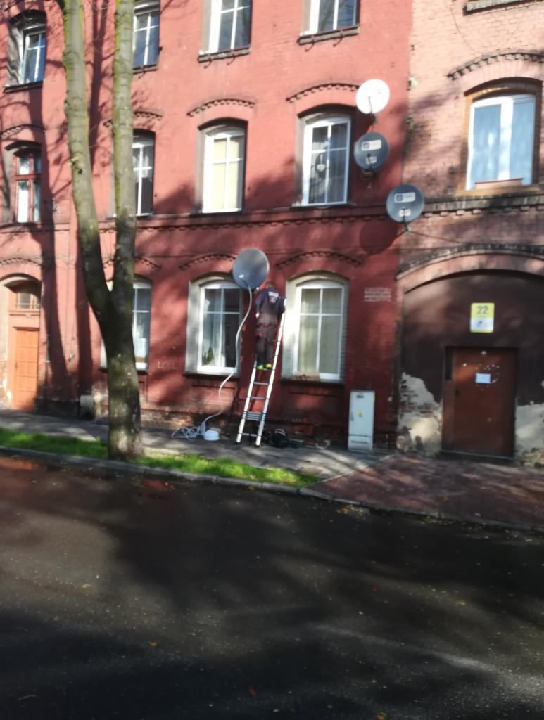 Instalator na drabinie montuje antenę satelitarną na ceglanym budynku, widoczne kable i narzędzia na chodniku, kilka anten już zamontowanych na elewacji.