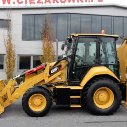 Żółta koparko-ładowarka Caterpillar 428F2 na tle budynku z szarymi oknami i drzewami, widok z boku.