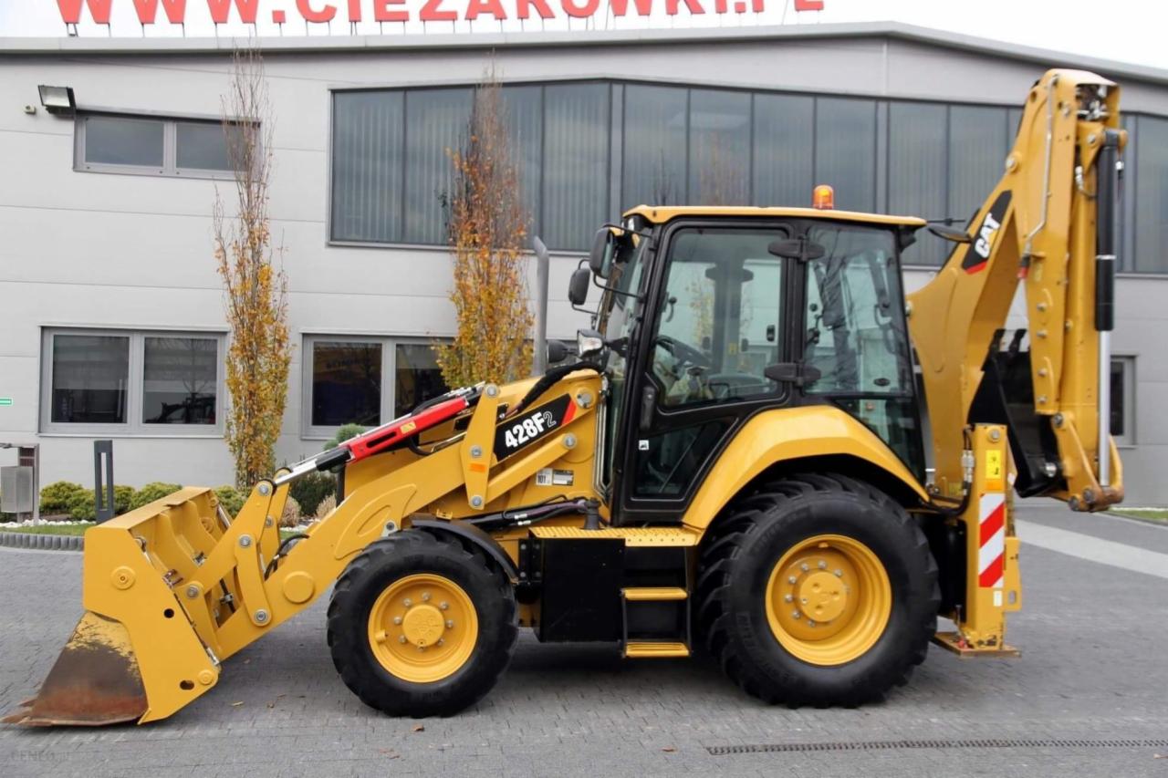 Żółta koparko-ładowarka Caterpillar 428F2 na tle budynku z szarymi oknami i drzewami, widok z boku.
