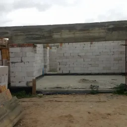 Wznoszenie ścian z bloczków betonowych na fundamencie z izolacją, widoczne rusztowania i drewniane palety z materiałami budowlanymi, surowy betonowy nadproże.