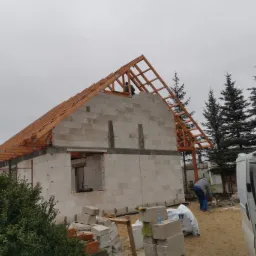 Wznoszenie ścian domu z bloczków betonu komórkowego z widoczną konstrukcją więźby dachowej, pracownik na dachu, teren budowy z zaparkowanym busem i stosem materiałów budowlanych.