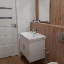 Nowoczesna łazienka z białą umywalką nablatową na szafce z szufladami, lustrem z podświetleniem LED i drewnianą okładziną ściany z widocznym przyciskiem spłuczki.