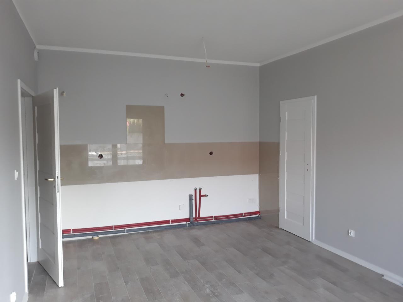 Wnętrze remontowanej kuchni z częściowo położoną glazurą, widocznymi instalacjami hydraulicznymi w kolorze czerwonym i szarymi panelami podłogowymi.