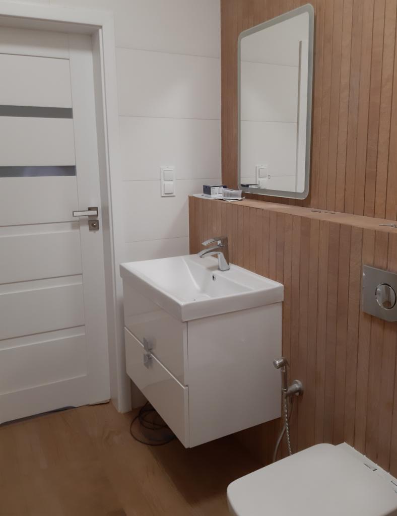 Nowoczesna łazienka z białą umywalką nablatową na szafce z szufladami, lustrem z podświetleniem LED i drewnianą okładziną ściany z widocznym przyciskiem spłuczki.