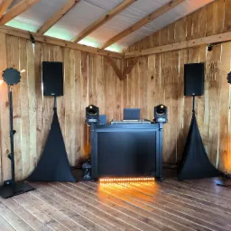 Scena z nagłośnieniem, oświetleniem i stanowiskiem DJ-a w drewnianym pawilonie, przygotowana na imprezę. Widoczne głośniki, reflektory, laptop i dekoracyjne lampy z żarówkami.