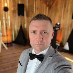 MAR-SOUND - Mężczyzna w szarej marynarce i muszce na tle drewnianej sceny z nagłośnieniem i oświetleniem.