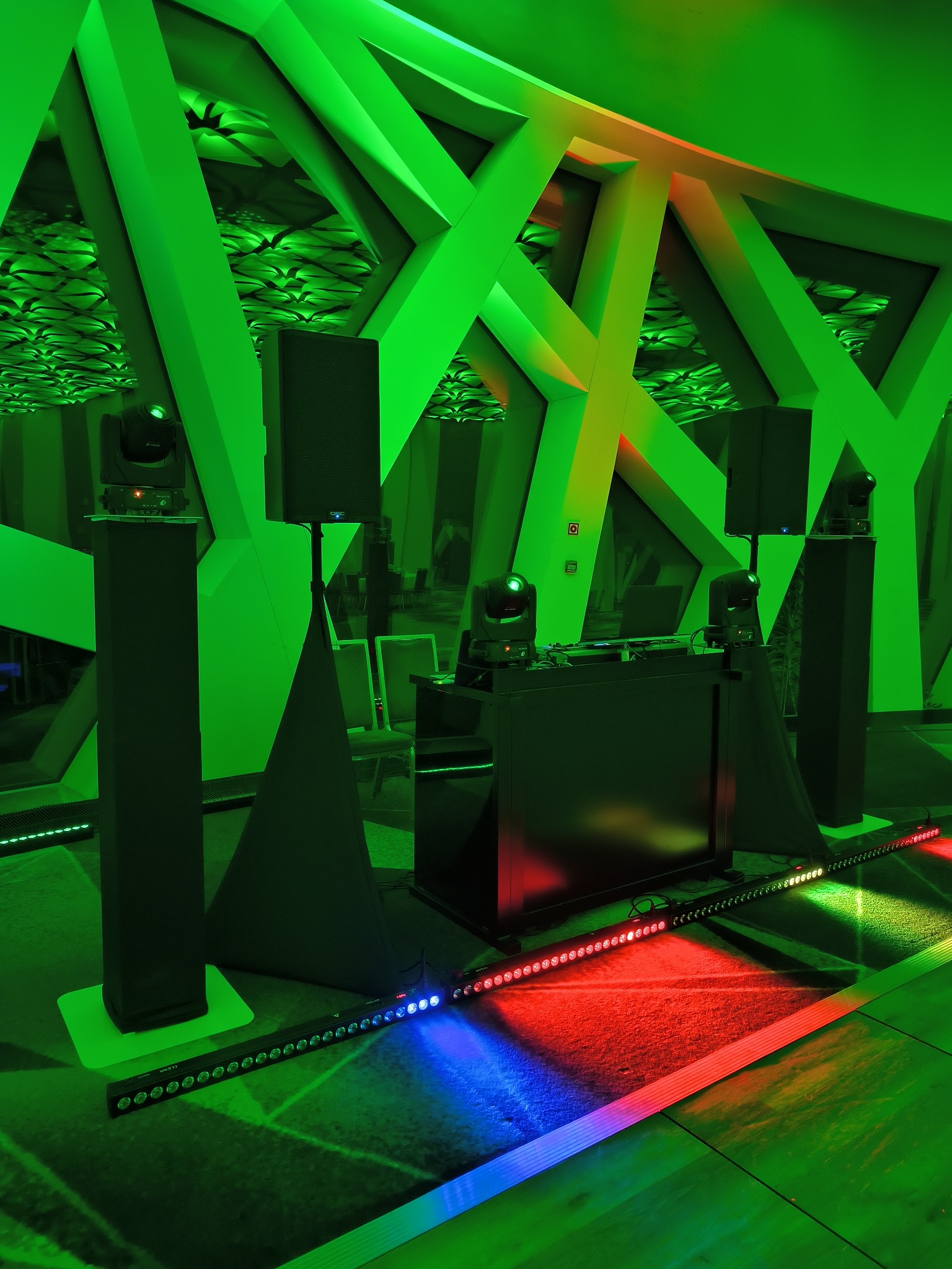 Scena z nagłośnieniem i oświetleniem w zielonej tonacji, widoczne kolumny głośnikowe, reflektory na statywach, konsoleta DJ-ska oraz listwa LED z kolorowymi światłami na podłodze, dekoracyjne...