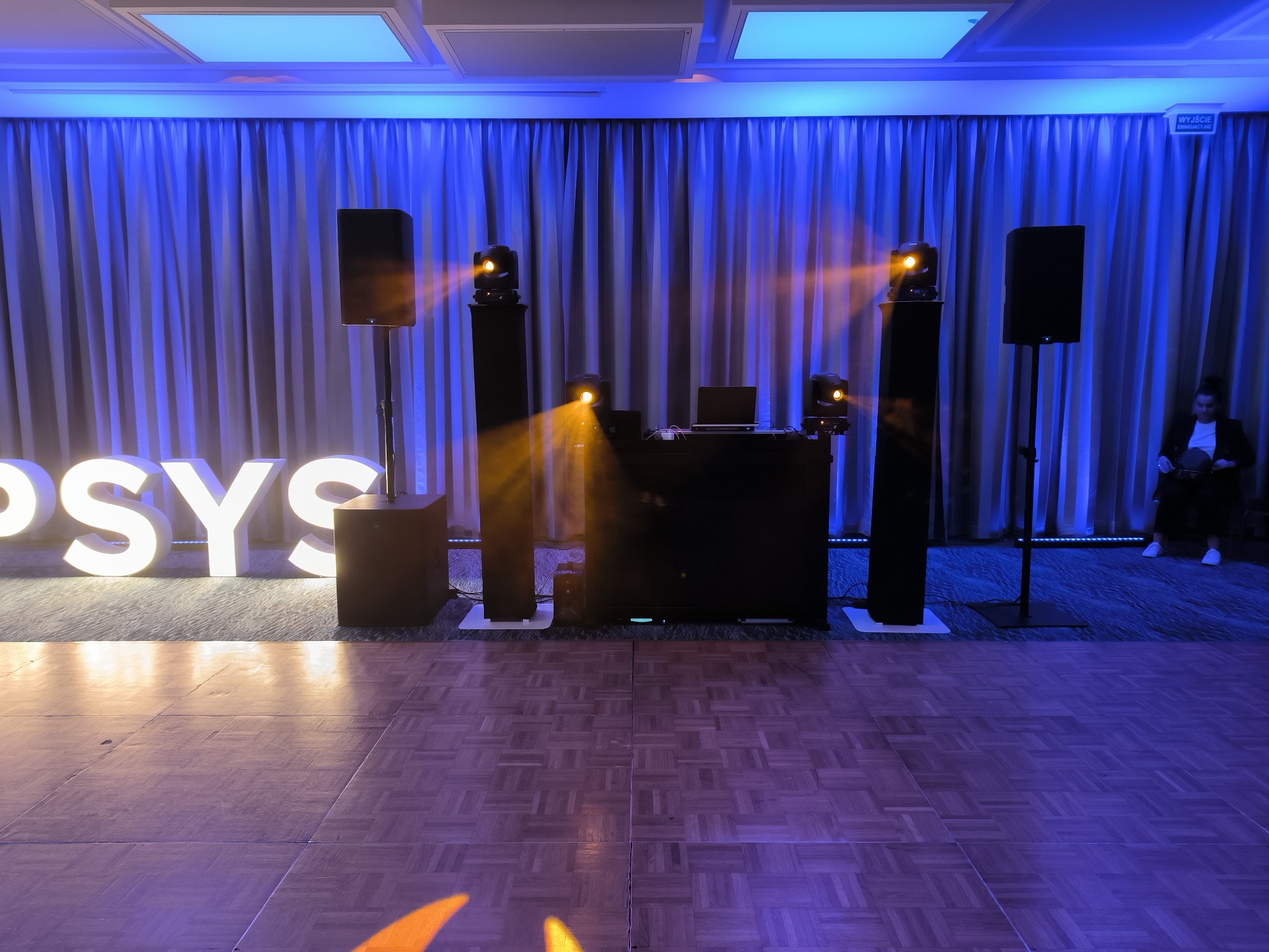 Parkiet taneczny oświetlony reflektorami, w tle kurtyna z niebieskim podświetleniem, kolumny głośnikowe, konsoleta DJ-ska z laptopem i podświetlany napis 'PSYS'.