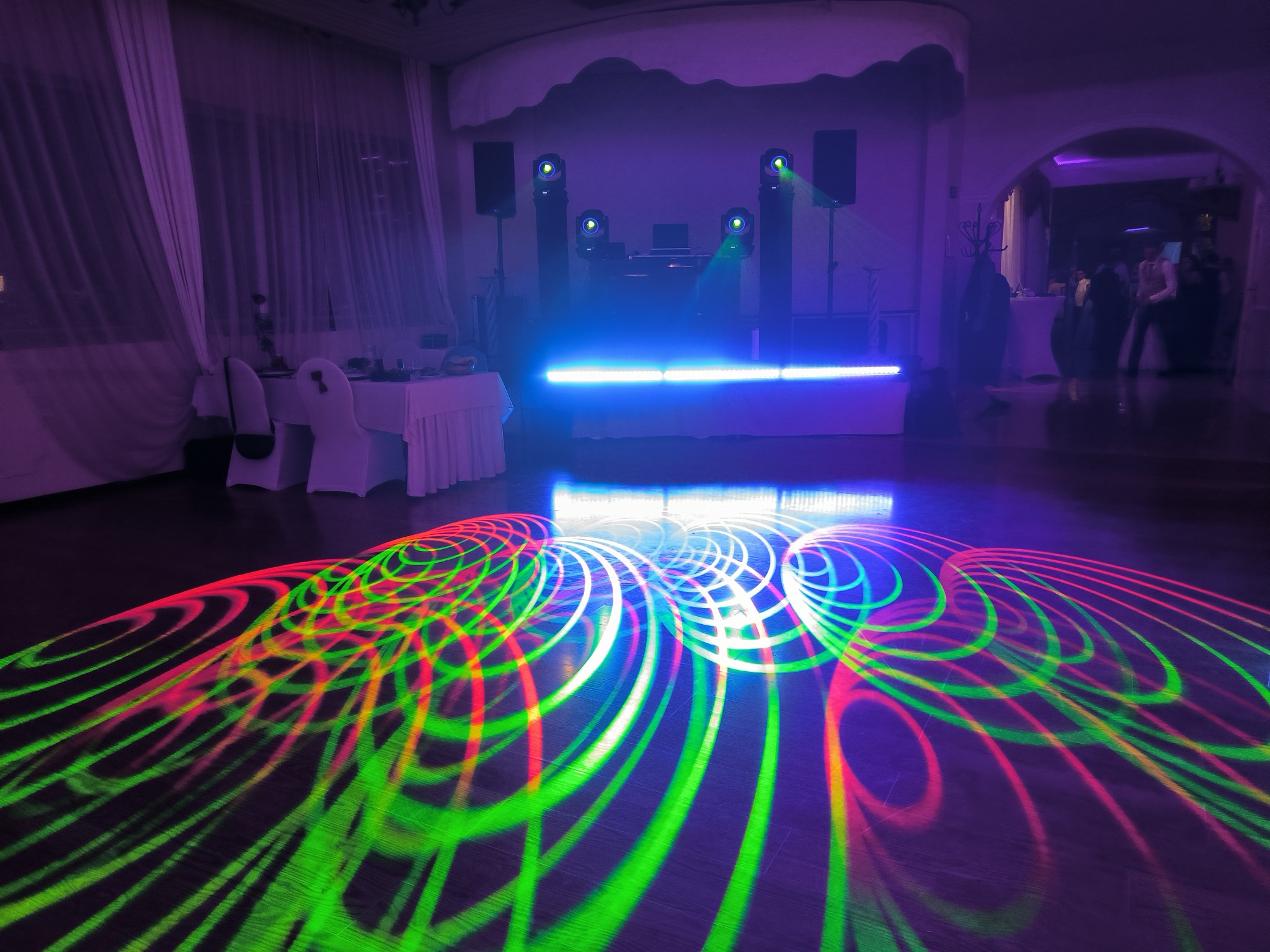 Sala balowa z oświetleniem dyskotekowym, na parkiecie widoczne kolorowe, okrągłe wzory świetlne, w tle scena z nagłośnieniem i didżejką.