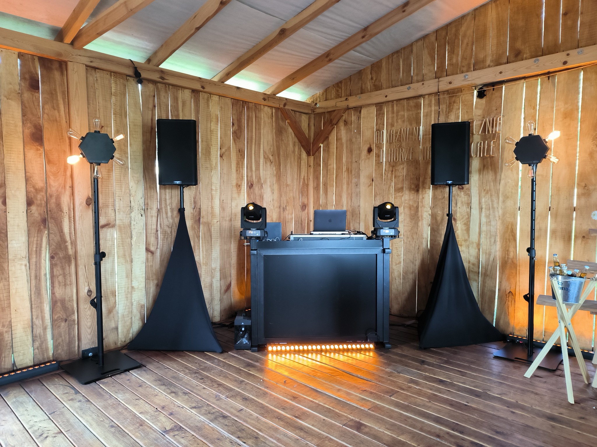 Scena z nagłośnieniem, oświetleniem i stanowiskiem DJ-a w drewnianym pawilonie, przygotowana na imprezę. Widoczne głośniki, reflektory, laptop i dekoracyjne lampy z żarówkami.