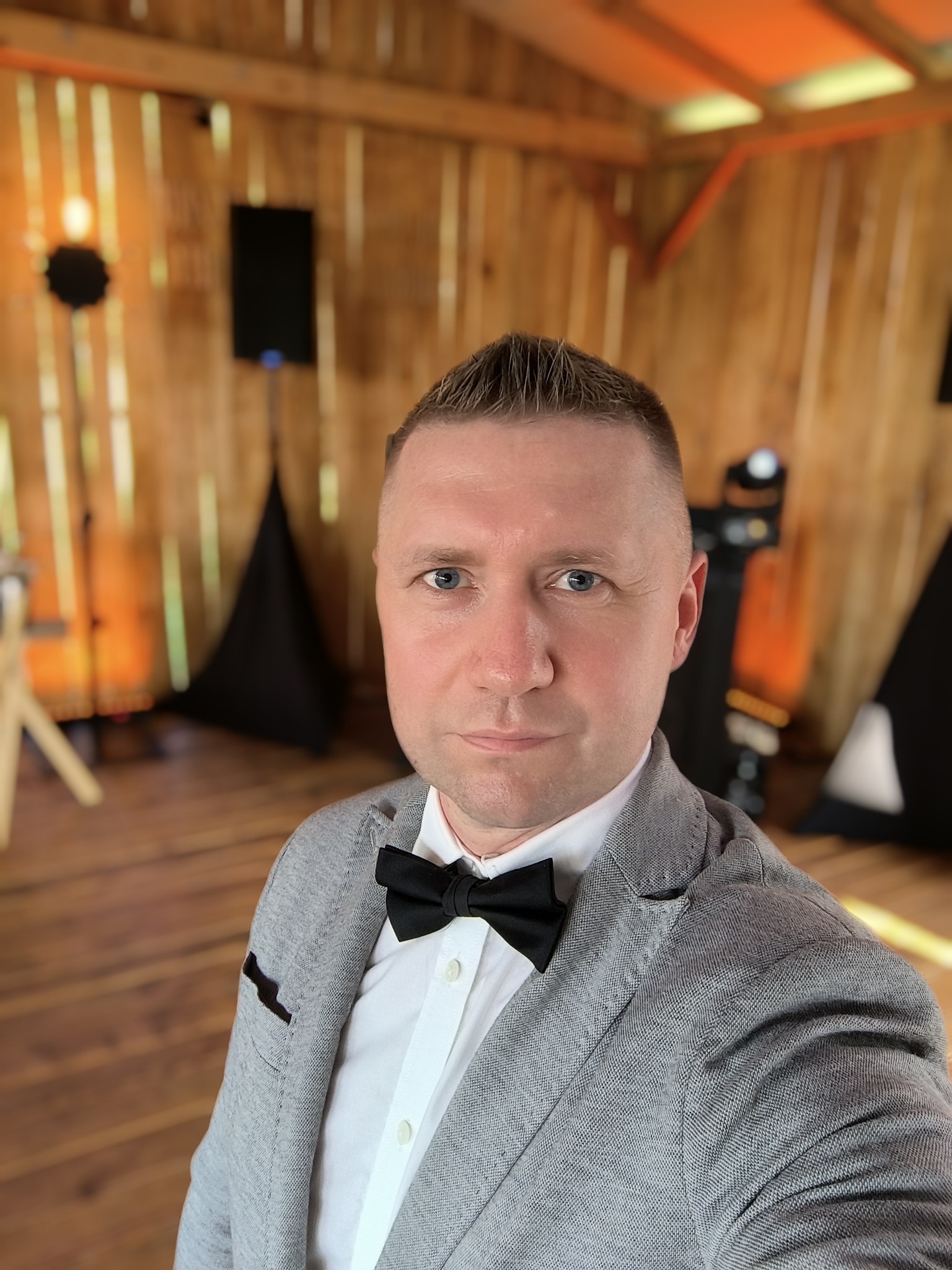 Mężczyzna w szarej marynarce i muszce na tle drewnianej sceny z nagłośnieniem i oświetleniem.