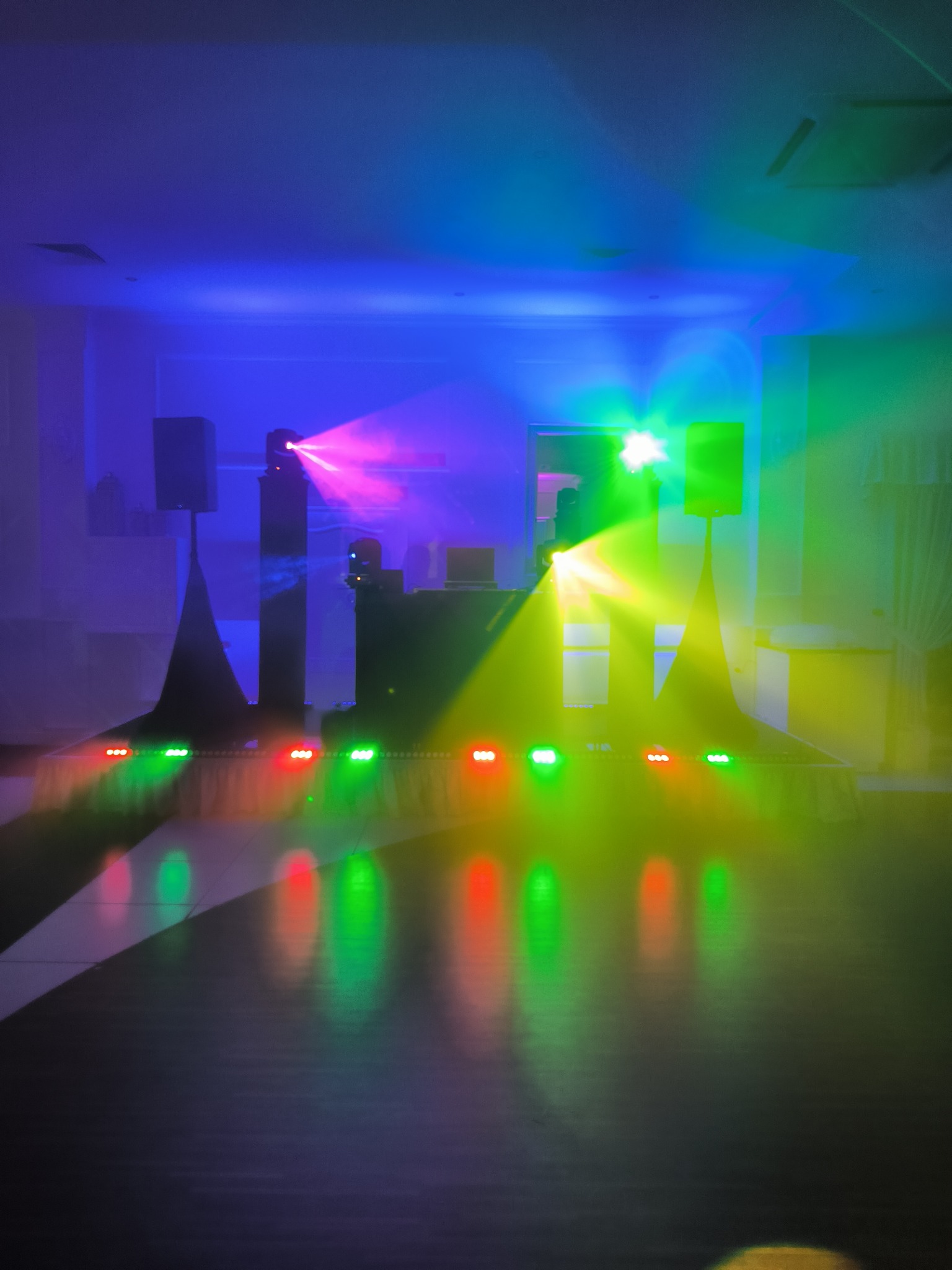 Scena oświetlona kolorowymi światłami, widoczne głośniki i sprzęt DJ-ski, tworzące dynamiczną atmosferę.