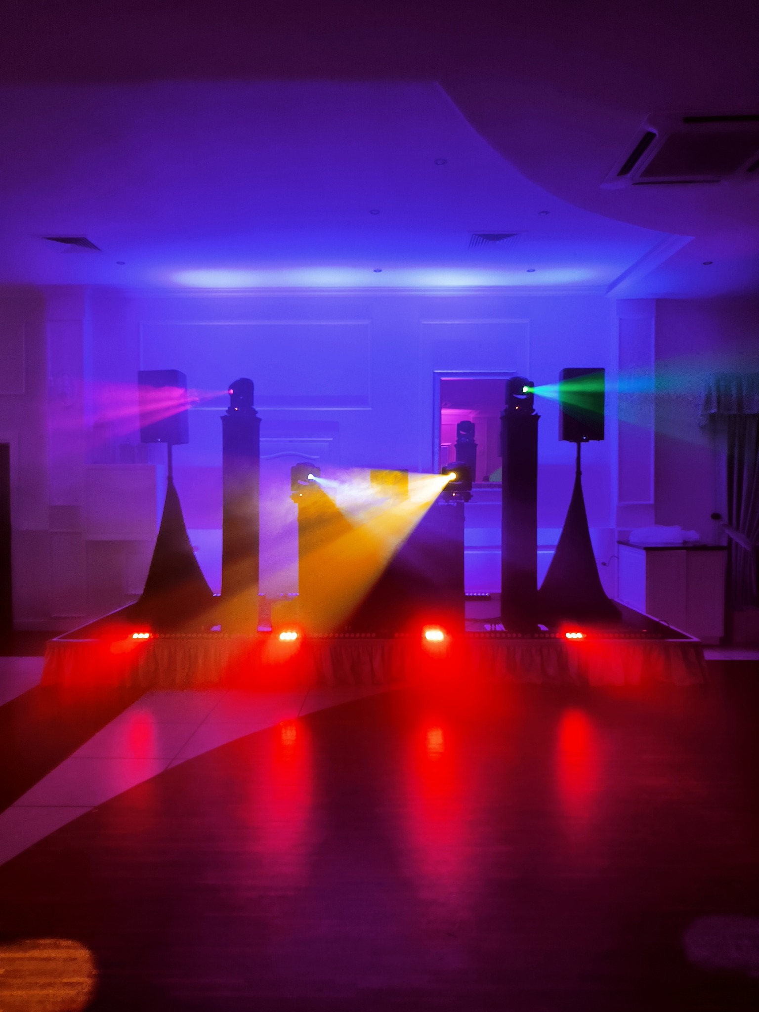 Scena oświetlona kolorowymi światłami, widoczne kolumny głośnikowe i reflektory, podświetlona podłoga parkietu.