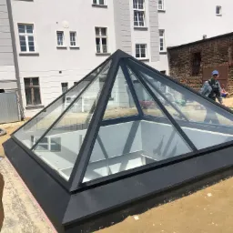 Piramidalne przeszklenie dachowe z czarną ramą, widoczne odbicie budynku w szklanych panelach, pracownik w tle.