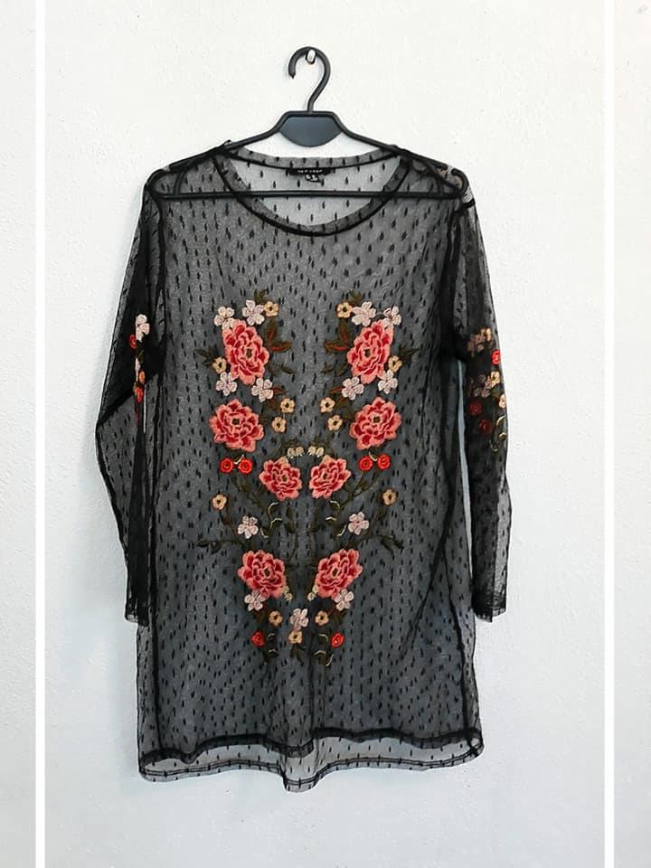 Czarna, przezroczysta tunika z długim rękawem, pokryta drobnymi kropkami i haftowanymi, symetrycznymi motywami kwiatowymi w odcieniach różu i pomarańczy, wisząca na czarnym wieszaku na tle białej...