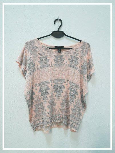 Różowo-szara bluzka z etnicznym wzorem zawieszona na czarnym wieszaku na tle jasnej ściany.