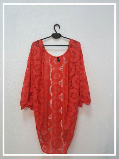 Pomarańczowa, ażurowa narzutka z okrągłym wzorem, wisząca na czarnym wieszaku na tle białej ściany.