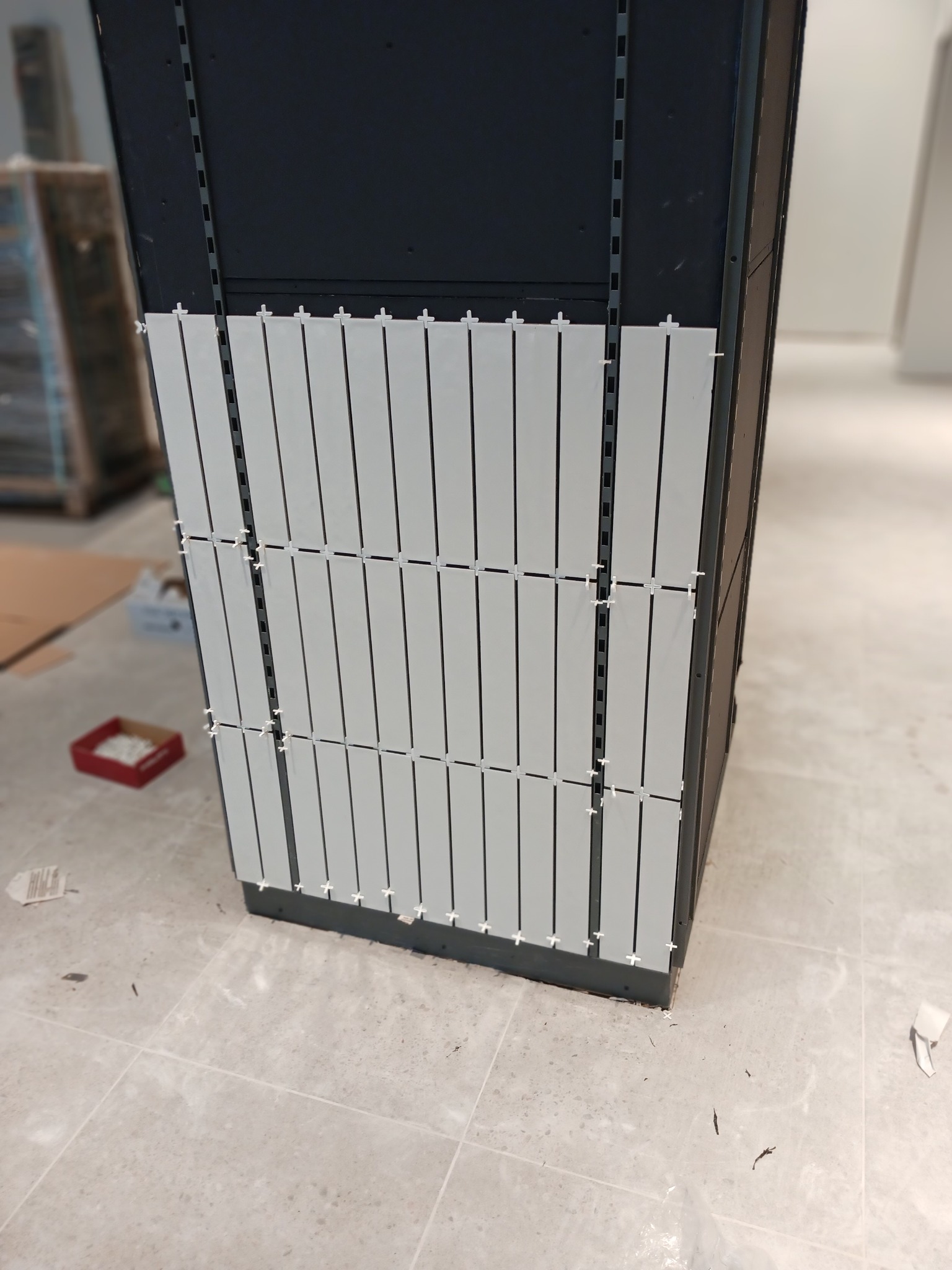 Montaż podłużnych, jasnych płytek na ciemnej konstrukcji z użyciem krzyżyków dystansowych, widoczna podłoga z jasnych płytek imitujących marmur.