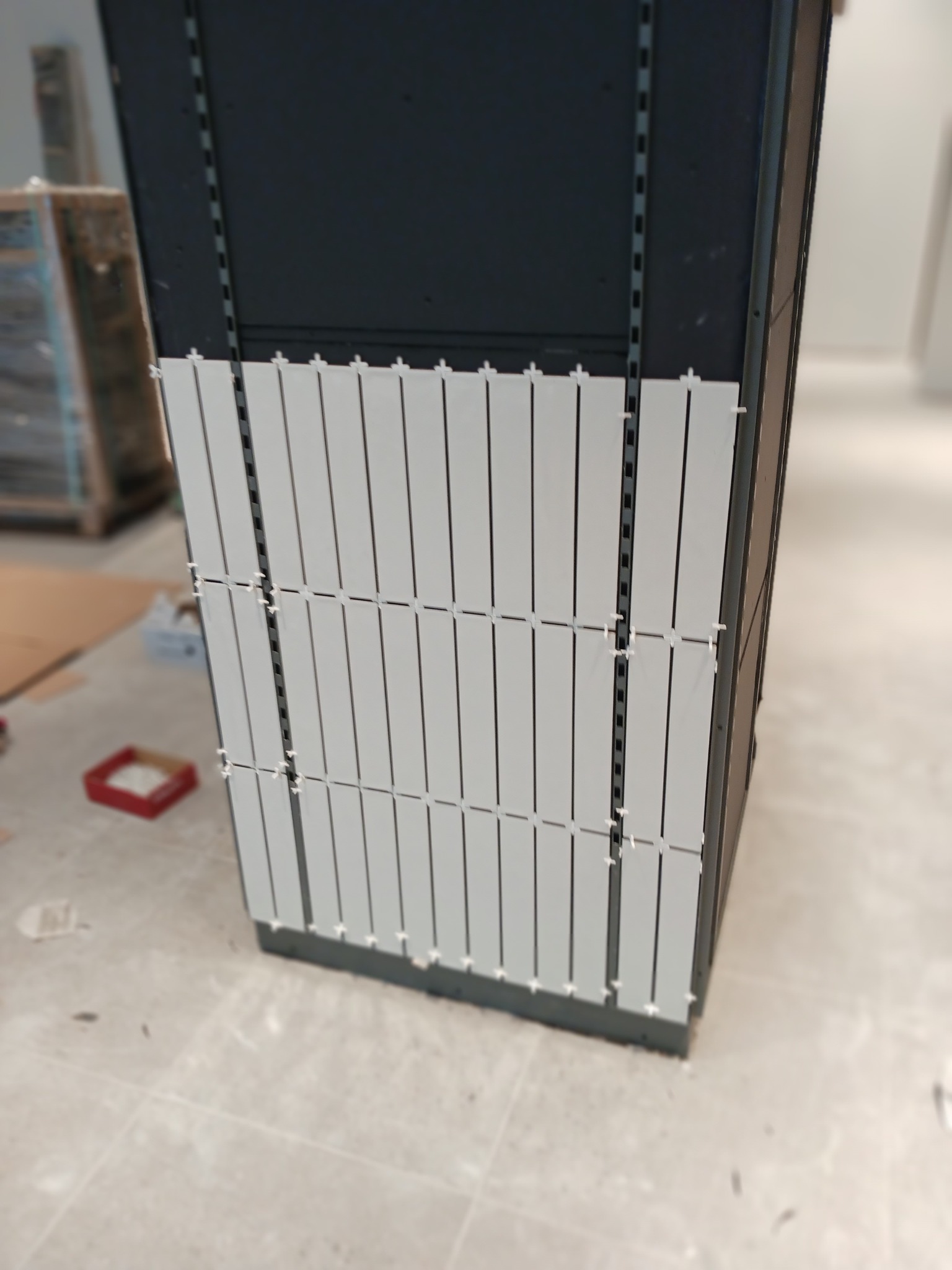 Montaż szarych, wąskich płytek na pionowej powierzchni z użyciem krzyżyków dystansowych, widoczne elementy konstrukcyjne i podłoże w trakcie prac wykończeniowych.