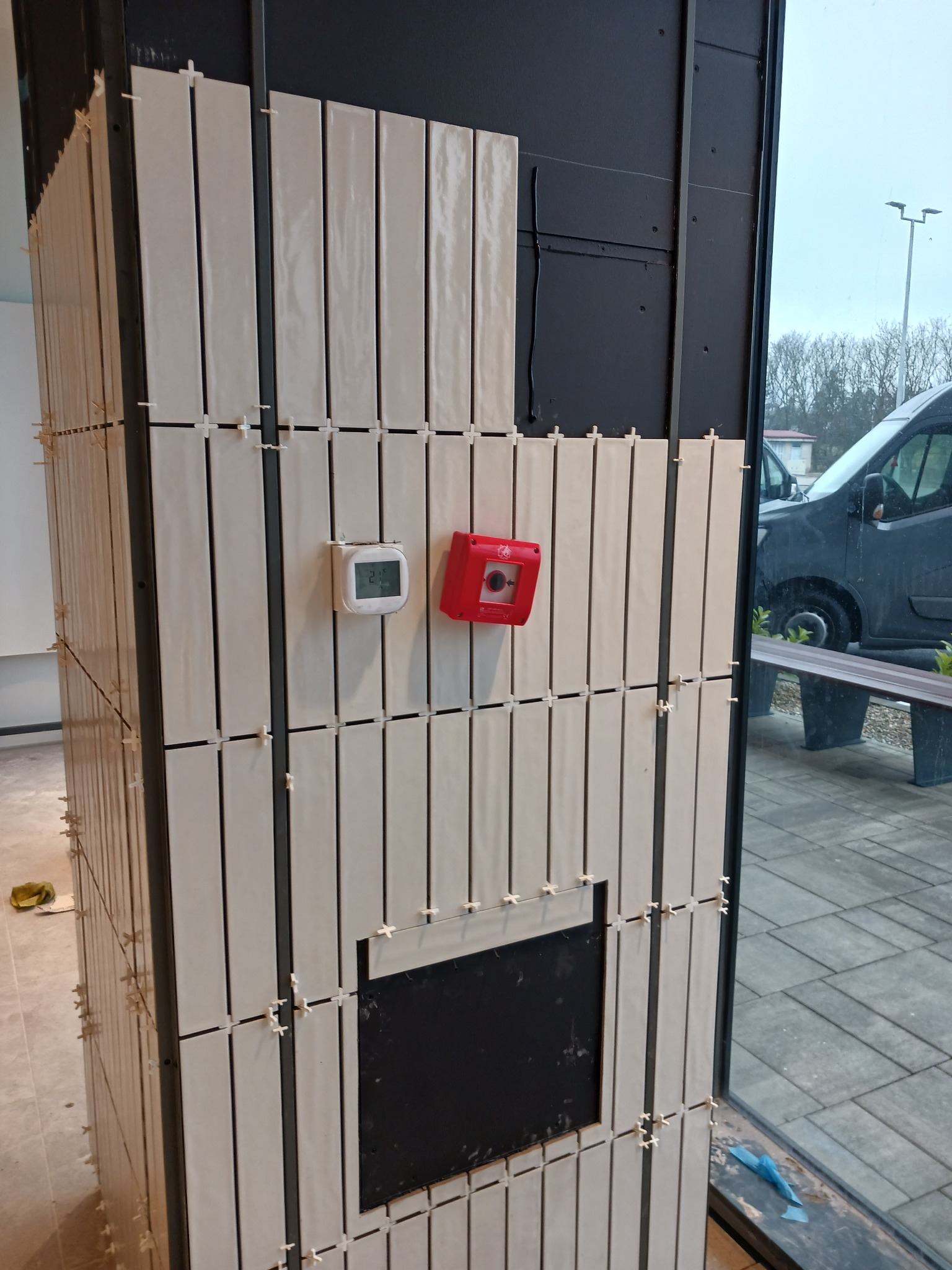 Kładzenie beżowych, błyszczących, podłużnych płytek na ścianie z widocznymi krzyżykami dystansowymi, termostatem i czujką przeciwpożarową