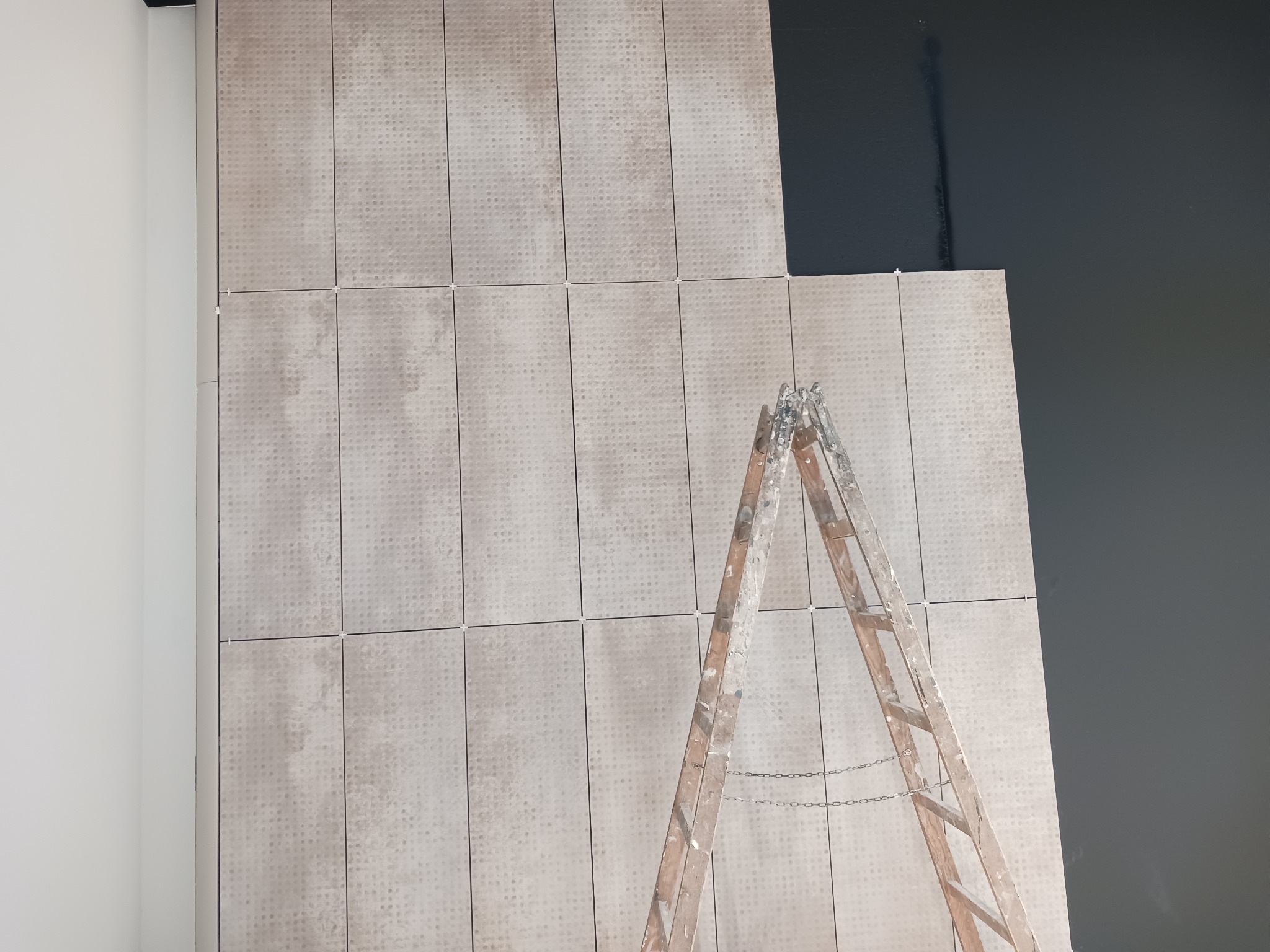 Ściana w trakcie wykańczania z beżowych, wzorzystych płytek ceramicznych, częściowo położonych, widoczna drabina malarska i fragment pomalowanej na ciemno ściany.