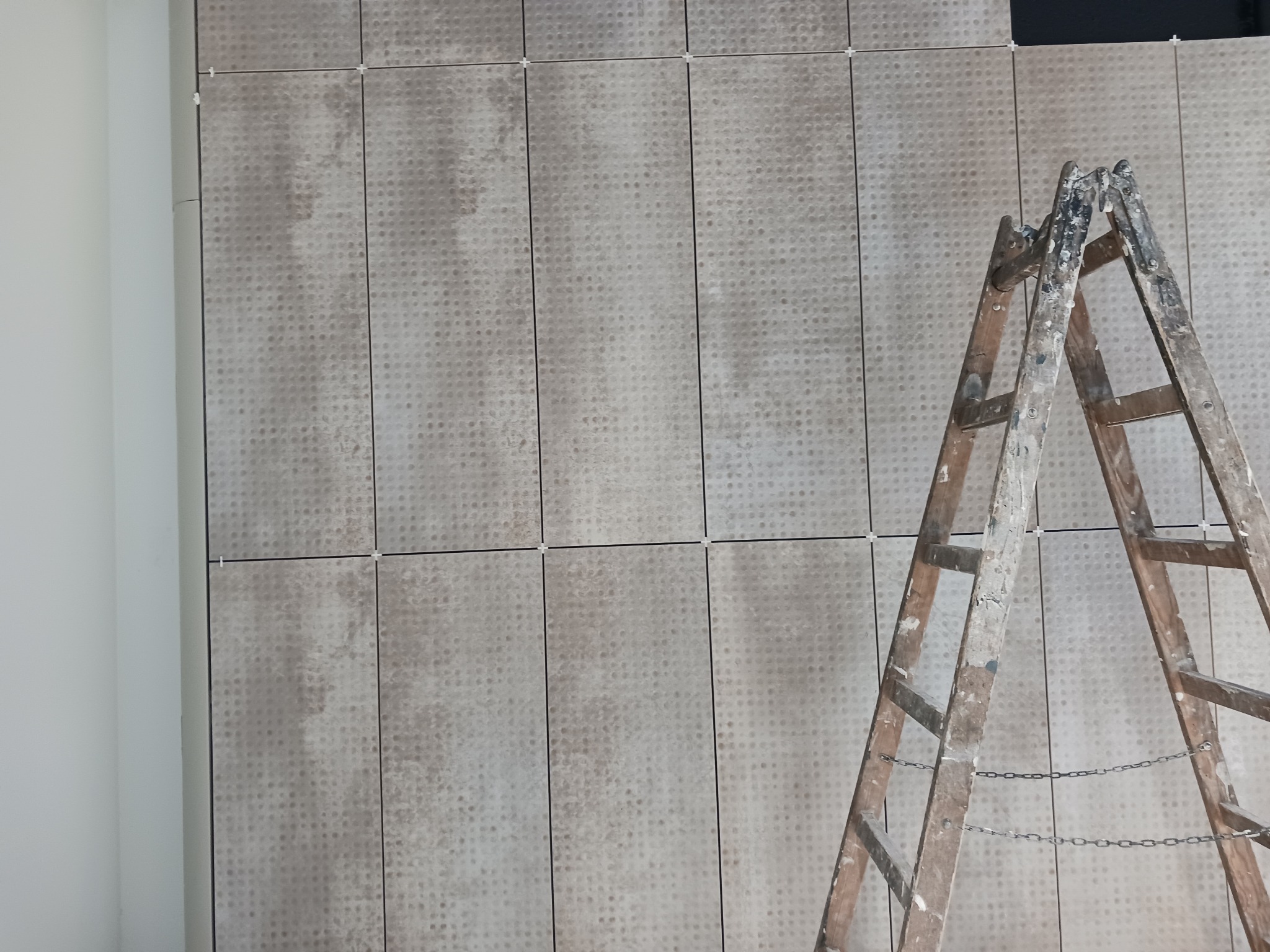 Ściana wyłożona beżowymi płytkami z delikatnym wzorem, widoczna drewniana drabina malarska z łańcuchem zabezpieczającym, białe krzyżyki dystansowe między płytkami.