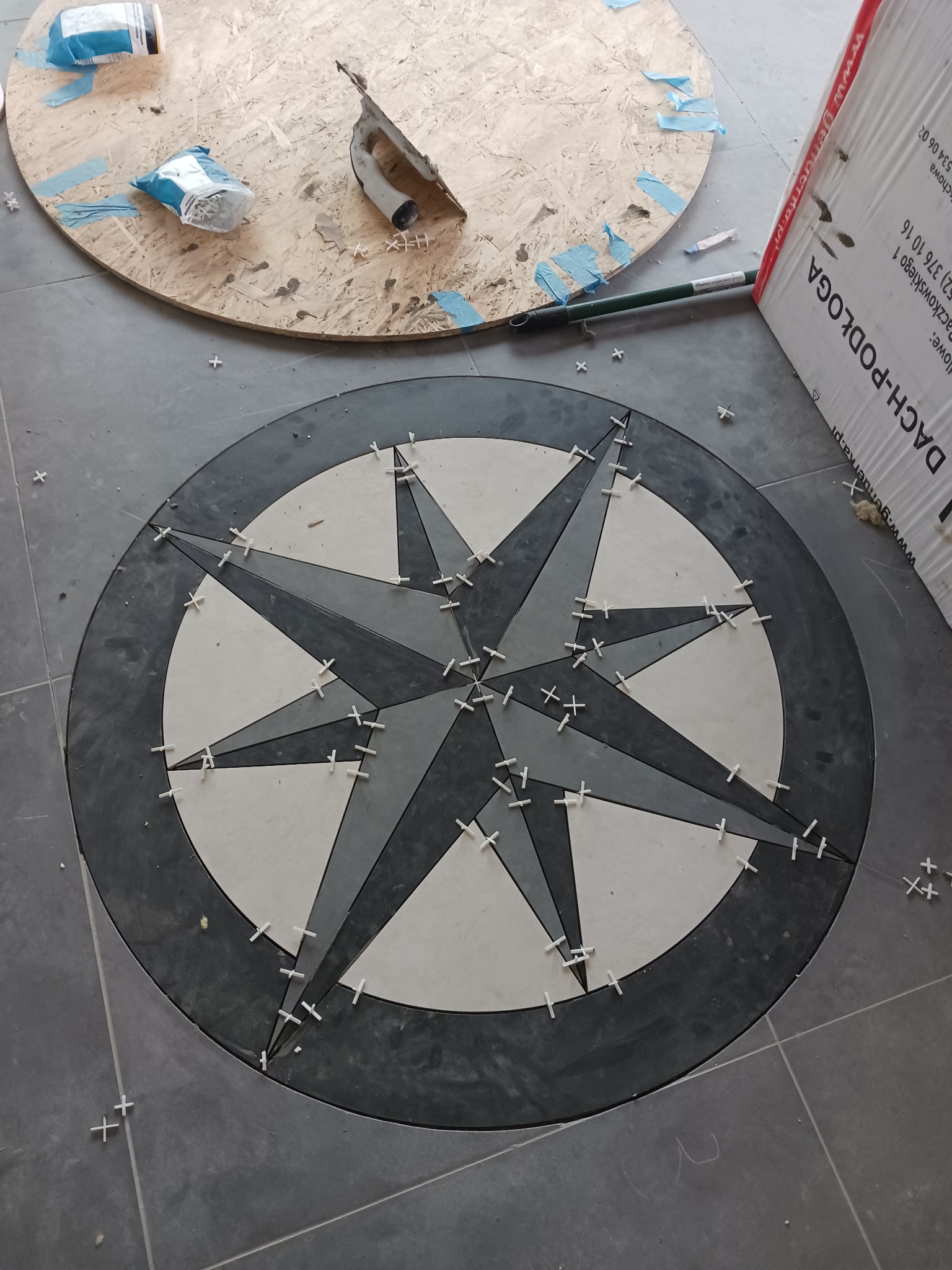 Układanie mozaiki podłogowej w kształcie rozety kompasu, z użyciem białych krzyżyków dystansowych, widoczne narzędzia i materiały na płycie OSB w tle.