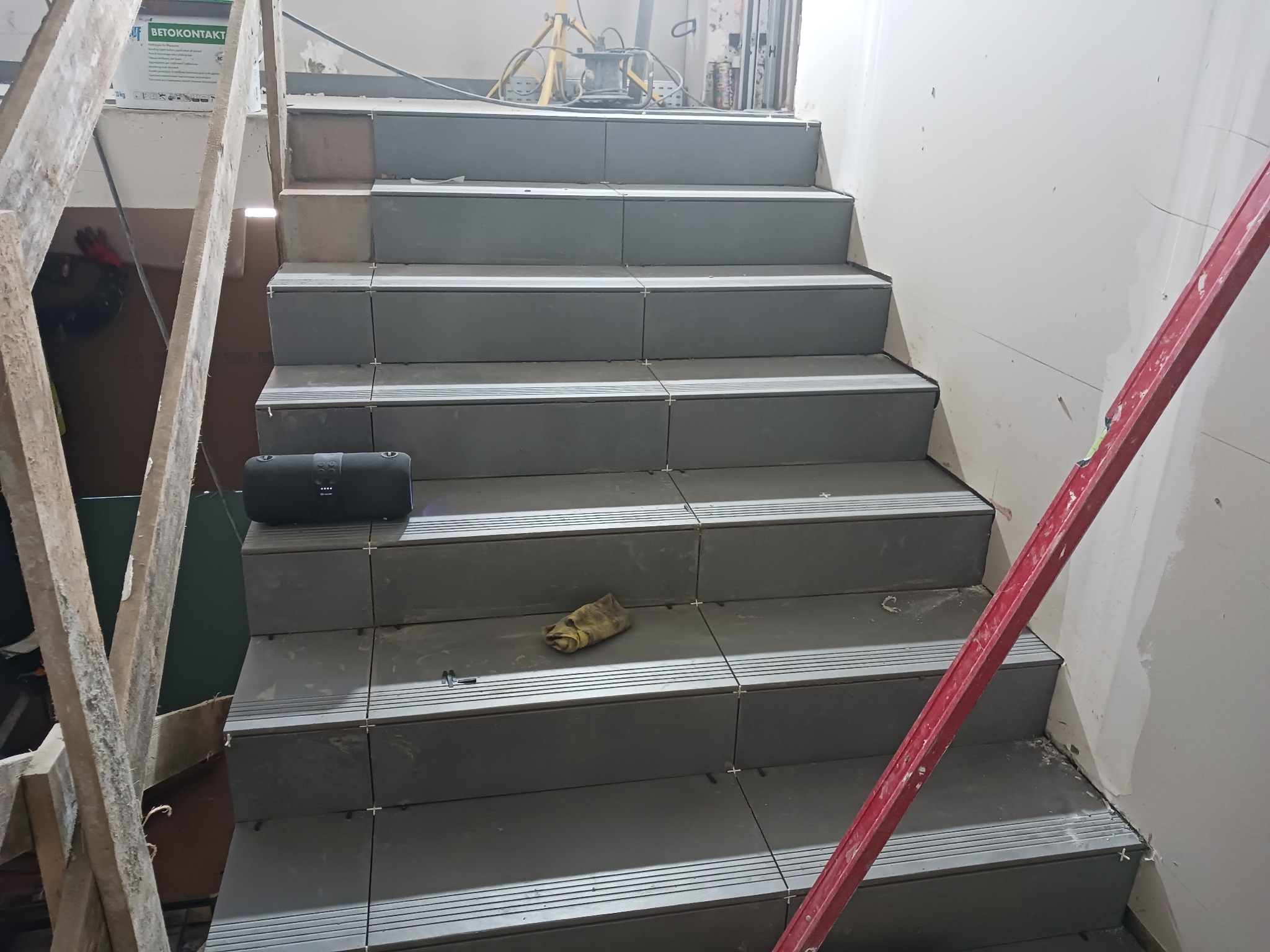 Schody wyłożone szarymi płytkami, widoczne krzyżyki dystansowe, poziomica i głośnik bluetooth na jednym ze stopni.