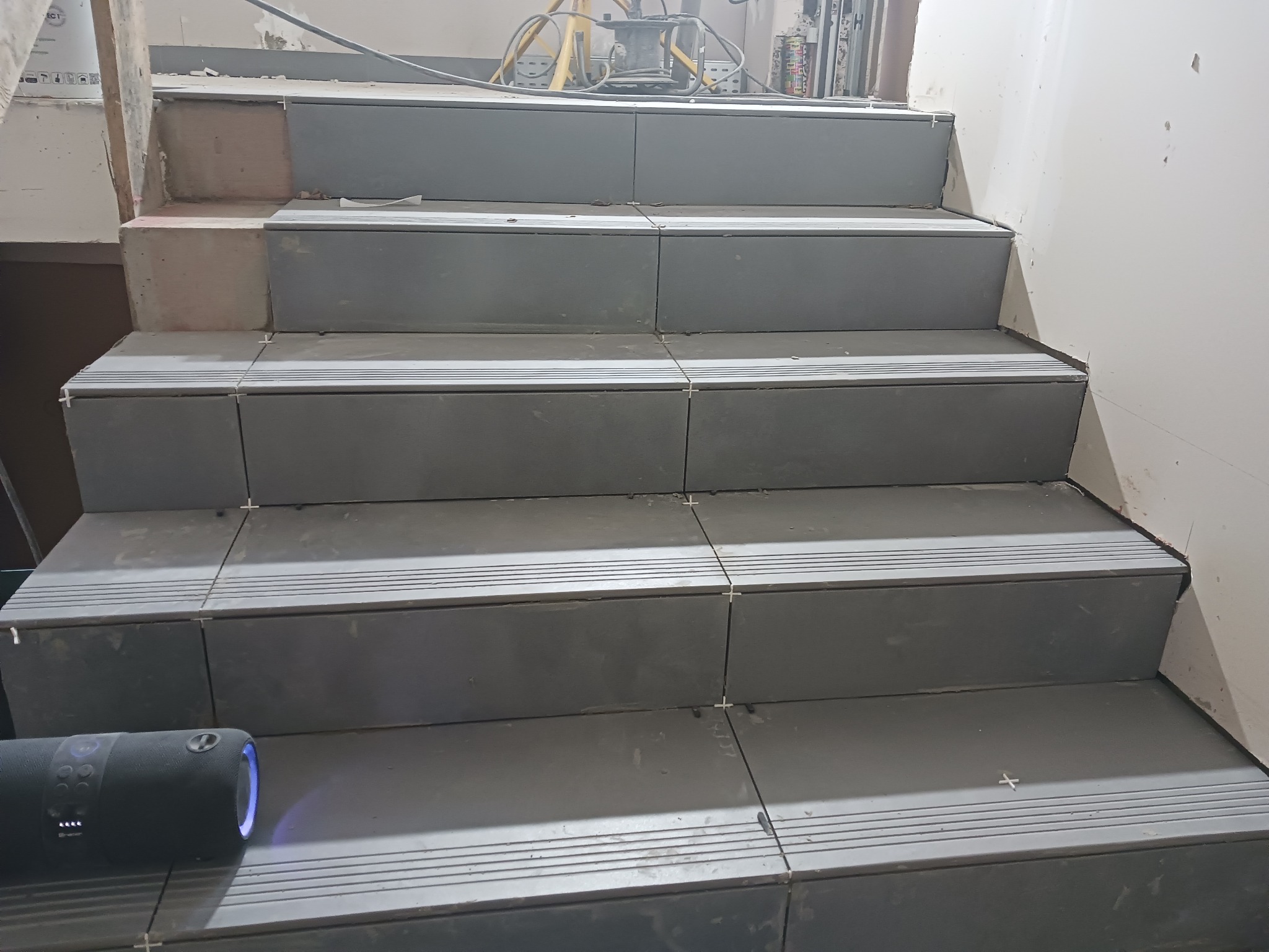 Schody w trakcie wykańczania z szarymi płytkami, widoczne krzyżyki dystansowe i głośnik przenośny na dole kadru.