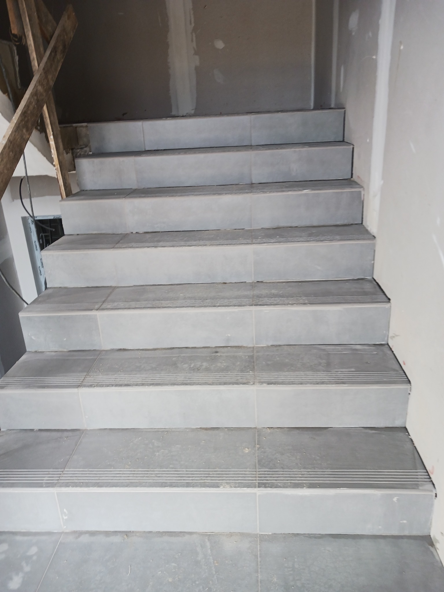 Wykończone schody wewnętrzne wyłożone szarymi płytkami z antypoślizgowymi rowkami, widoczne niedoskonałości wykończenia przy ścianie i drewniany element konstrukcyjny w górnym lewym rogu.