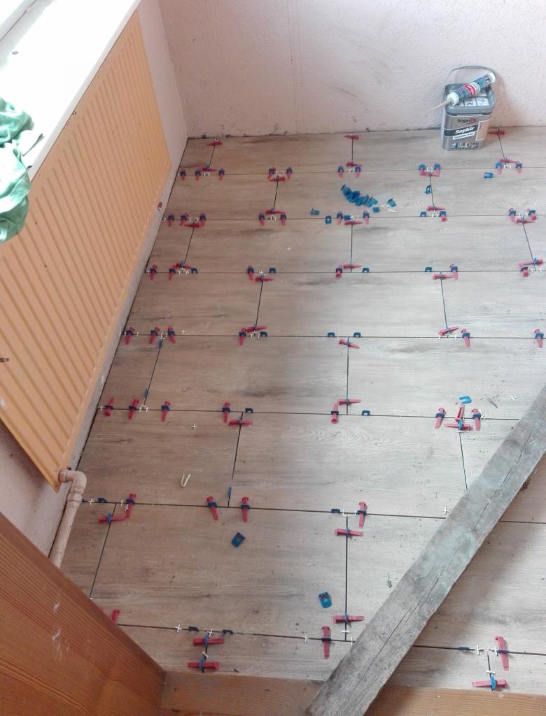 Ułożone płytki podłogowe imitujące drewno, z widocznymi krzyżykami dystansowymi i klinami poziomującymi, w tle grzejnik żeberkowy i wiadro z klejem.