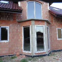 Nowe białe okna PCV w surowym stanie budynku z czerwonej cegły, widoczne ślady pianki montażowej i niezakończone prace wykończeniowe, na dachu widoczne okna połaciowe.