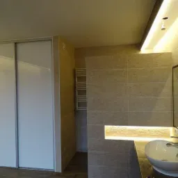 Nowoczesna łazienka z beżowymi płytkami, podświetlanym lustrem, białą umywalką i szafą z przesuwnymi drzwiami.