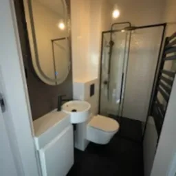 Nowoczesna, wąska łazienka z czarną podłogą i białymi elementami. Umywalka narożna z czarną armaturą, owalne lustro z podświetleniem LED, wisząca toaleta oraz kabina prysznicowa z czarnymi ramami.