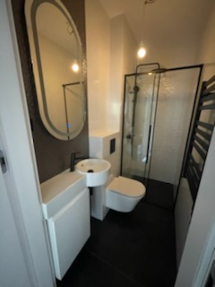 Nowoczesna, wąska łazienka z czarną podłogą i białymi elementami. Umywalka narożna z czarną armaturą, owalne lustro z podświetleniem LED, wisząca toaleta oraz kabina prysznicowa z czarnymi ramami.