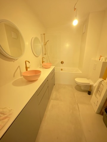 Nowoczesna łazienka w trakcie wykańczania: różowe umywalki nablatowe z mosiężną armaturą, biała wanna i toaleta, szara szafka pod umywalki, okrągłe lustra. Widoczne elementy wykończeniowe...