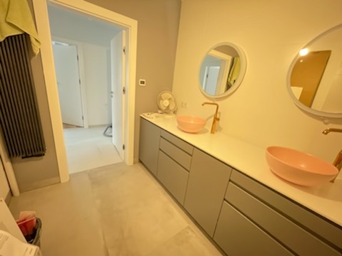 Wykończona łazienka z dwoma okrągłymi lustrami, różowymi umywalkami nablatowymi i szarą zabudową szafkową; widoczny grzejnik drabinkowy i wentylator na blacie.