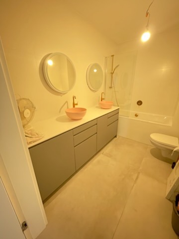 Wykończona łazienka z dwoma okrągłymi lustrami, różowymi umywalkami nablatowymi i szarą szafką pod umywalką, widoczna wanna z parawanem i toaleta.