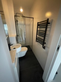 Wąska łazienka z czarnymi akcentami: kabina prysznicowa z czarną ramą, wisząca toaleta, umywalka nablatowa na białej szafce, czarny grzejnik drabinkowy i ciemna podłoga.