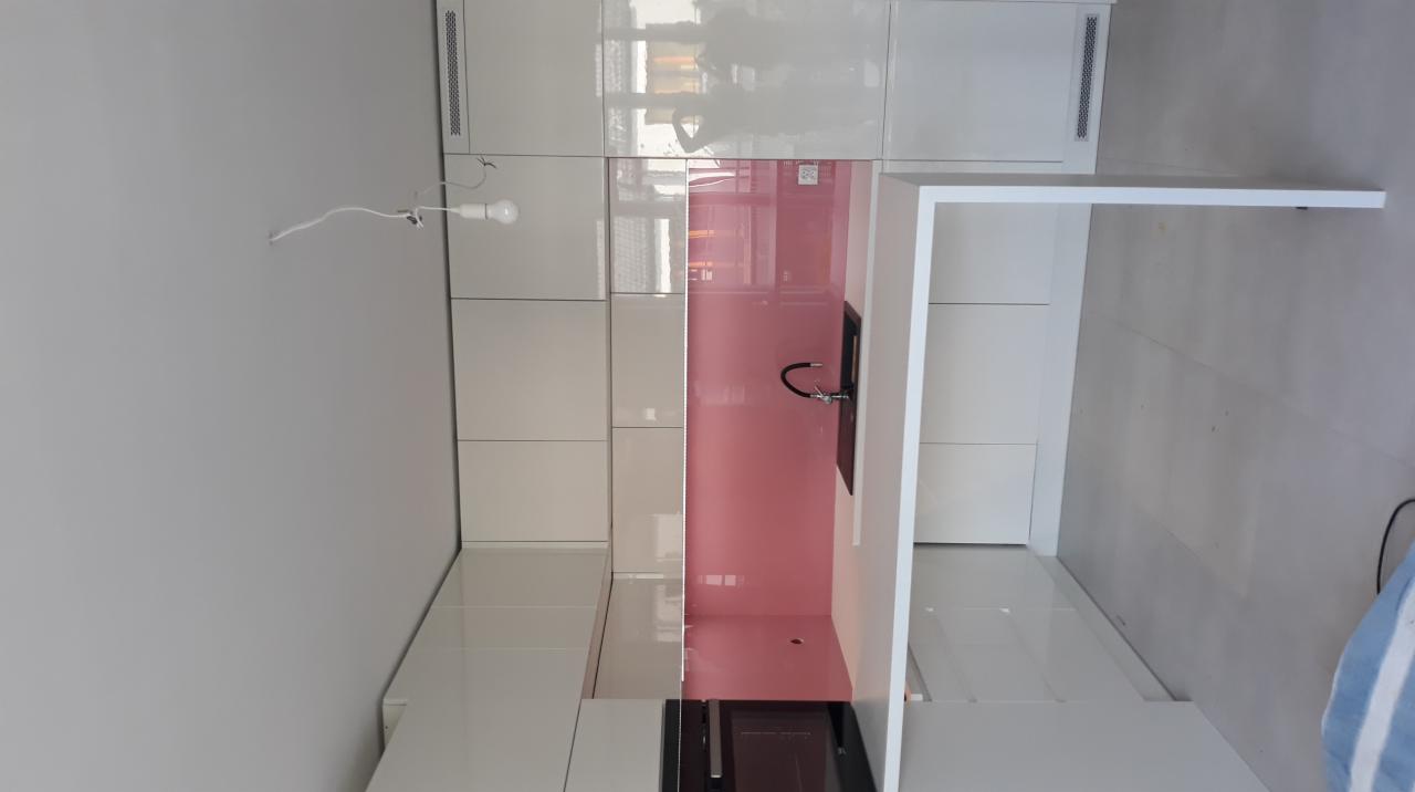 Nowoczesna, biała kuchnia z różowym panelem szklanym nad blatem i minimalistyczną armaturą, oświetlona pojedynczą żarówką na kablu.